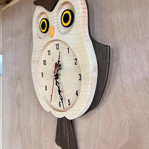 Puede incluir: Un reloj de madera con forma de b&uacute;ho con cuerpo marr&oacute;n y cara blanca. El reloj tiene agujas negras y ojos amarillos. Los n&uacute;meros del 1 al 12 est&aacute;n impresos en la esfera del reloj.