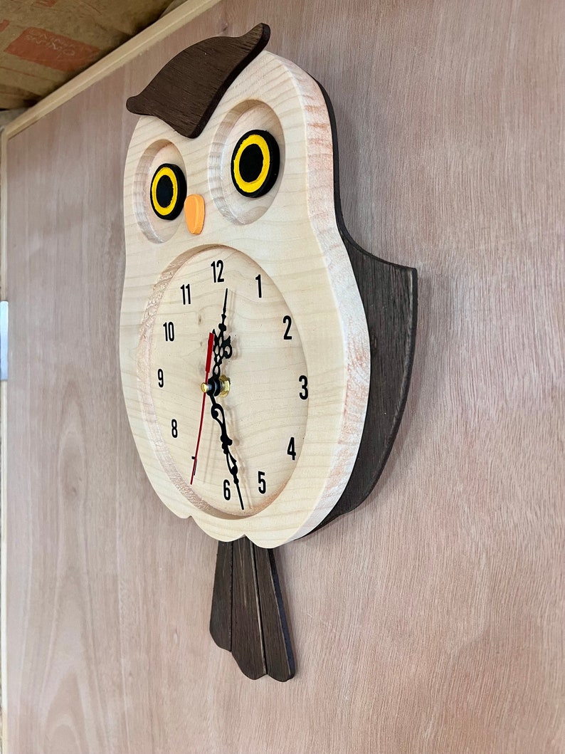 Puede incluir: Un reloj de madera con forma de b&uacute;ho con cuerpo marr&oacute;n y cara blanca. El reloj tiene agujas negras y ojos amarillos. Los n&uacute;meros del 1 al 12 est&aacute;n impresos en la esfera del reloj.