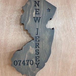 May include: A wooden wall hanging in the shape of New Jersey, stained in a dark gray color. The state's name and zip code 07470 are vertically carved into the wood. A small star is carved near the top.