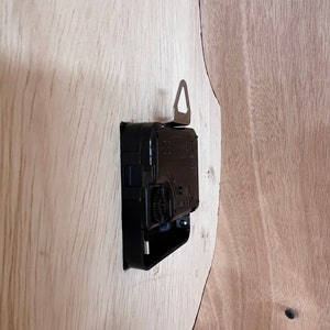 Puede incluir: Un mecanismo de reloj negro con un gancho de suspensi&oacute;n plateado fijado a una superficie de madera marr&oacute;n claro.