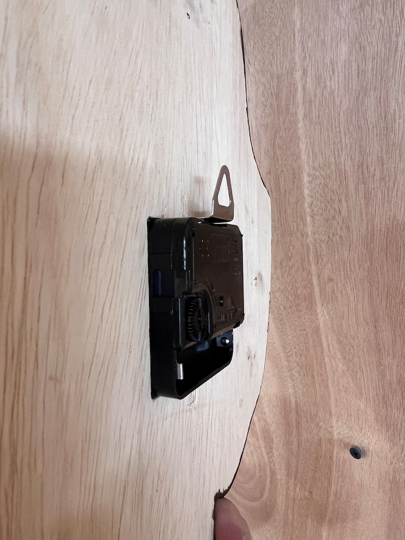 Puede incluir: Un mecanismo de reloj negro con un gancho de suspensi&oacute;n plateado fijado a una superficie de madera marr&oacute;n claro.