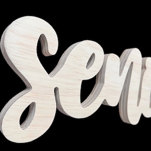 May include: A wooden sign that spells out the name "Semia" in a cursive font. The sign is made of light-colored wood and has a three-dimensional effect, with the letters slightly raised from the background. The background is black.