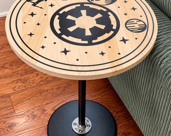 May include: A round side table with a light wood top featuring a black graphic print of a space station, a starfighter, a planet, and a central imperial emblem surrounded by stars. The table has a black metal pedestal base and a circular black foot.