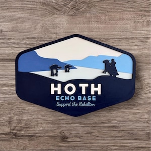 May include: A navy blue and white layered wooden sign with a hexagonal shape. The sign features a snowy landscape with AT-AT walkers and a silhouette of a person riding a Tauntaun. The text reads "HOTH ECHO BASE Support the Rebellion."
