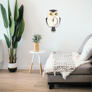 Puede incluir: Un reloj de madera con forma de b&uacute;ho con detalles en negro y marr&oacute;n cuelga de una pared blanca. La esfera del reloj tiene n&uacute;meros y manecillas negros. El b&uacute;ho tiene ojos amarillos. El reloj est&aacute; colgado sobre un sof&aacute; blanco con una manta y almohadas grises.