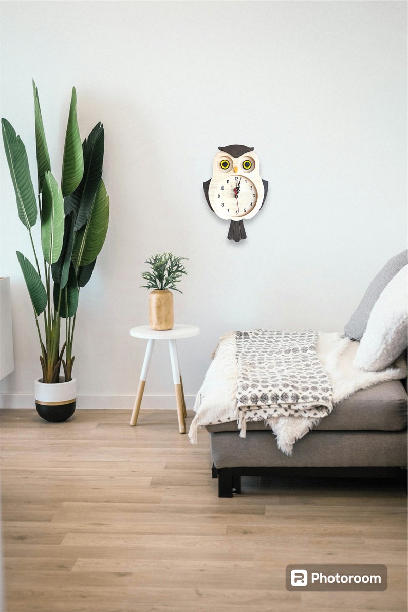 Puede incluir: Un reloj de madera con forma de b&uacute;ho con detalles en negro y marr&oacute;n cuelga de una pared blanca. La esfera del reloj tiene n&uacute;meros y manecillas negros. El b&uacute;ho tiene ojos amarillos. El reloj est&aacute; colgado sobre un sof&aacute; blanco con una manta y almohadas grises.