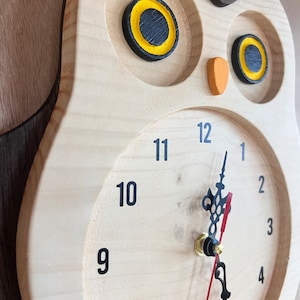 Puede incluir: Un reloj de madera con forma de b&uacute;ho con agujas negras y ojos amarillos. La esfera del reloj tiene n&uacute;meros negros del 1 al 12.