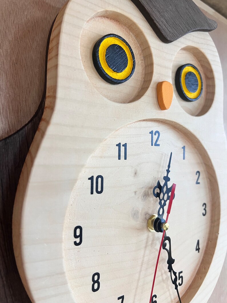 Puede incluir: Un reloj de madera con forma de b&uacute;ho con agujas negras y ojos amarillos. La esfera del reloj tiene n&uacute;meros negros del 1 al 12.