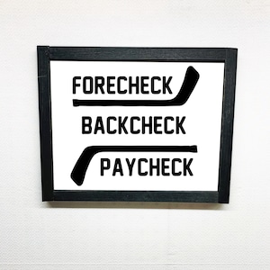 May include: Black and white sign with the words "Forecheck", "Backcheck", and "Paycheck" with hockey stick graphics.