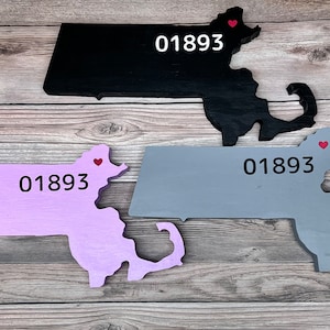 May include: Three wooden Massachusetts state cutouts in black, lavender, and gray. Each has the number "01893" printed on it in black, and a small red heart. The cutouts are displayed on a wooden background.