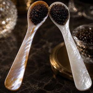 May include: Two mother-of-pearl caviar spoons filled with black caviar are positioned to touch. The spoons are on a dark surface, with a small glass bowl of caviar and champagne flutes in the background. The spoons have a pearlescent sheen.