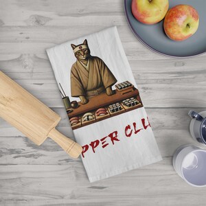 May include: A white kitchen towel featuring a cartoon cat wearing a red headband and a brown robe. The cat is standing behind a counter with sushi rolls and the text "Sushi Chef" in red.