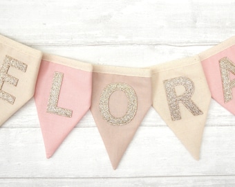 May include: A pink and beige fabric banner with the name "ELORA" spelled out in gold glitter letters.