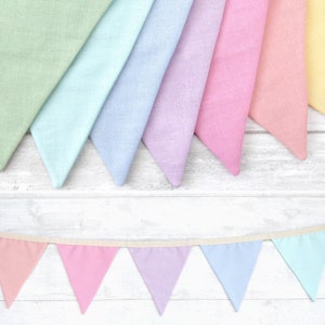 May include: A selection of pastel-coloured triangular flags, arranged in a row, with a matching banner below. The flags are in shades of green, blue, purple, pink, and yellow. The banner is strung on a cream-coloured ribbon, creating a cheerful, decorative display.