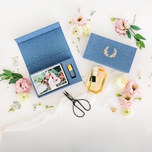 May include: A flat lay featuring a blue box with a photo, a gold-colored USB drive, and a smaller blue box with a gold wreath design. Pink flowers, greenery, and scissors are arranged on a white surface.