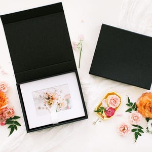 May include: A black presentation box with a white matted photograph inside, next to a closed box. The image is surrounded by orange and pink roses, anemones, and greenery. A small pair of scissors and a yellow dish with flowers are also present.