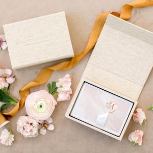 May include: A beige linen-textured box with a magnetic lid, containing a ring on a white cushion. A long mustard-yellow silk ribbon winds around the box, accompanied by several light pink ranunculus and sweet pea flowers scattered nearby.