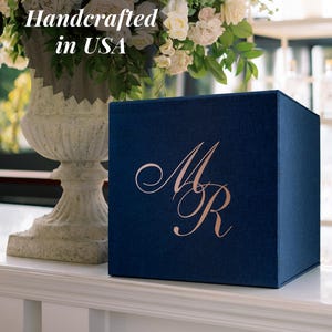 Può includere: Una scatola quadrata blu navy con un monogramma in oro rosa raffigurante le lettere M e R sulla parte anteriore. Il testo Handcrafted in USA è visibile nell'angolo in alto a sinistra, parzialmente oscurato da un grande bouquet di fiori bianchi.