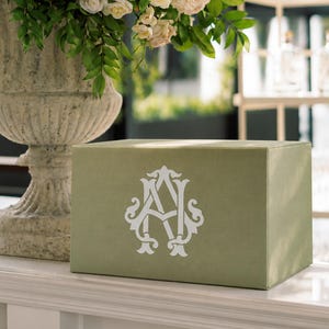May include: A rectangular sage green box featuring a white monogram of the letters A and N intertwined in a decorative script font. The box is positioned in front of a large stone urn filled with a dense arrangement of cream and white roses.