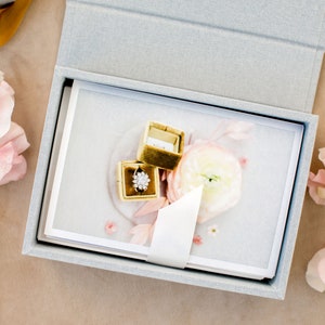 May include: A light grey rectangular keepsake box containing a stack of white cards with a floral print. A small gold velvet ring box sits on top, holding a silver ring with a floral-shaped cluster of clear stones, surrounded by pink flowers.
