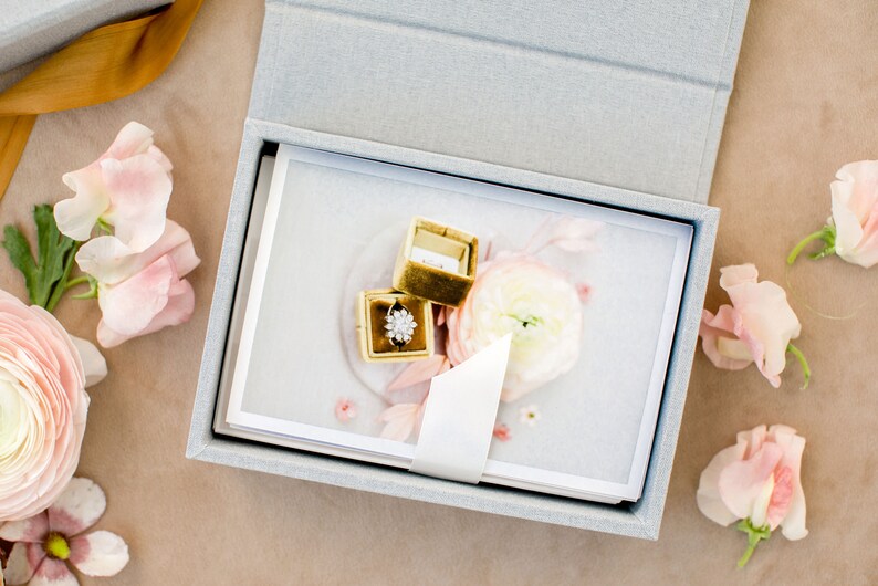 May include: A light grey rectangular keepsake box containing a stack of white cards with a floral print. A small gold velvet ring box sits on top, holding a silver ring with a floral-shaped cluster of clear stones, surrounded by pink flowers.