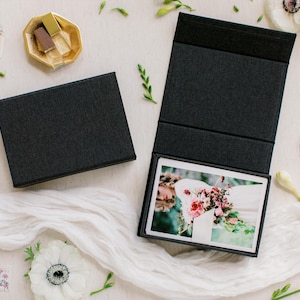 May include: Two black presentation boxes, one open to reveal a photograph of a floral arrangement. The boxes are on a white surface with white fabric, flowers, and small decorative items. The boxes are likely for photos or keepsakes.