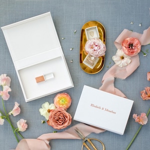 Op de afbeelding: Een witte rechthoekige doos met een houten USB-stick erin, een tweede witte doos met de tekst "Elisabeth & Johnathan" op het deksel, een gouden ovaal dienblad met een ringdoosje en een roze bloem, en roze lint met bloemstengels.