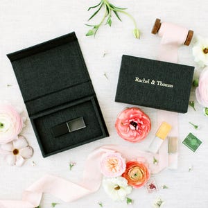 May include: Two dark charcoal gray textured boxes, one open and one closed, featuring the text Rachel & Thomas. The open box contains a black rectangular USB drive. The set is surrounded by light pink ribbon and various pastel-colored flowers.