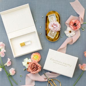 May include: An open white box with a gold and white object inside, next to a gold tray holding two ring boxes. A closed white box with gold text and a pair of scissors are also present. Pink flowers and ribbon are arranged on a blue-grey surface.