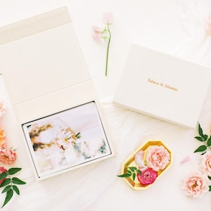 May include: A cream-coloured rectangular photo box with a magnetic lid, featuring the gold embossed text "Rebecca & Sebastian" on the cover. The open box reveals a stack of photographs, accompanied by a small gold tray holding pink flowers.