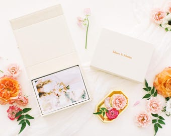 May include: A cream-colored rectangular photo box with a magnetic lid, featuring the gold embossed text "Rebecca & Sebastian" on the cover. The open box reveals a stack of photographs, accompanied by a small gold tray holding pink flowers.