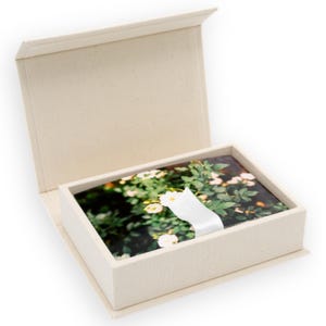 May include: A rectangular, off-white, hinged storage box containing a stack of rectangular cards. The cards feature a photographic print of green foliage with small white flowers. A white ribbon is wrapped around the center of the card stack.