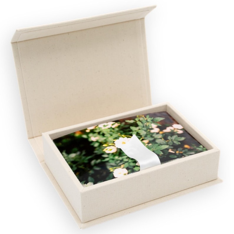 May include: A rectangular, off-white, hinged storage box containing a stack of rectangular cards. The cards feature a photographic print of green foliage with small white flowers. A white ribbon is wrapped around the center of the card stack.
