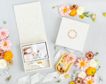 Puede incluir: Un set de cajas de color crema con textura de lino que incluye una tapa cuadrada con un emblema de corona de color cobre, una caja abierta con tarjetas atadas con una cinta blanca y una pequeña bandeja dorada con dos viales de vidrio, rodeado de dalias.