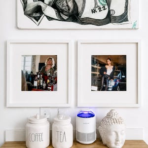 May include: Two white square frames containing photographs, one showing a person at a bar and the other a person by a car. Below are two white ceramic canisters labeled COFFEE and TEA, a white KATCHY insect trap, and a white Buddha head sculpture.