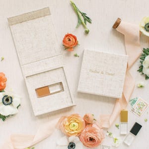 Op de afbeelding: Een vierkante, getextureerde, gebroken witte doos met de tekst Pauline & Daniel op het deksel, vergezeld van een open bijpassende doos, perzik- en witte bloemen, een lichtroze lint en twee kleine glazen flesjes met gouden en zwarte doppen.
