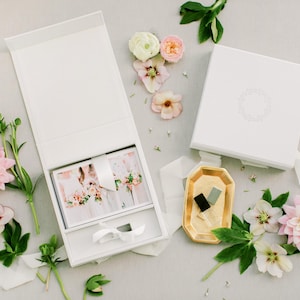 May include: A white presentation box, open to reveal a photo album with a ribbon tie. The box is surrounded by pink and white flowers, greenery, and a gold-colored tray with small items. The overall aesthetic is soft and elegant.