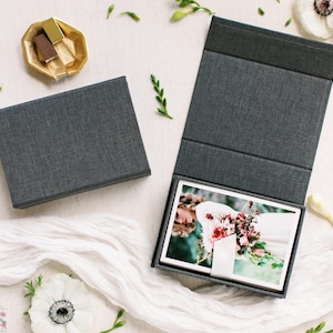 May include: Two dark gray fabric-covered boxes, one closed and one open, containing a stack of rectangular photographic prints. The open box features a hinged lid and a white ribbon pull tab, displaying a photo of a person holding a floral bouquet.