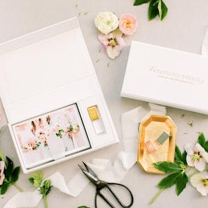 May include: White gift boxes with gold lettering and floral arrangements. One box is open, revealing a photo and a small perfume bottle. A gold tray holds nail polish. Scissors and ribbon are also present.