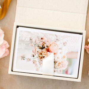 May include: A rectangular, cream-colored fabric-covered box containing a stack of white-bordered photographic prints. The top print displays a floral arrangement featuring soft pink and peach-toned flowers. A white ribbon secures the stack of prints.