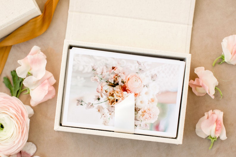 May include: A rectangular, cream-colored fabric-covered box containing a stack of white-bordered photographic prints. The top print displays a floral arrangement featuring soft pink and peach-toned flowers. A white ribbon secures the stack of prints.