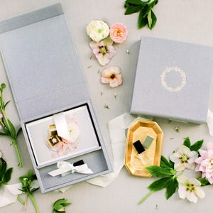 Op de afbeelding: Een lichtgrijze rechthoekige geschenkdoos met een magnetisch deksel, zowel open als gesloten afgebeeld. De open doos bevat een witte kaart met een lint en een klein item. Een goudkleurig achthoekig dienblad bevat twee kleine rechthoekige objecten.