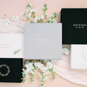 May include: A collection of five square and rectangular boxes in black, white, and light gray with fabric-like textures. Some boxes feature gold or silver foil text including names like Aaron & Lauren, Jacob & LeeAnna, and Michael & Joanna with dates.