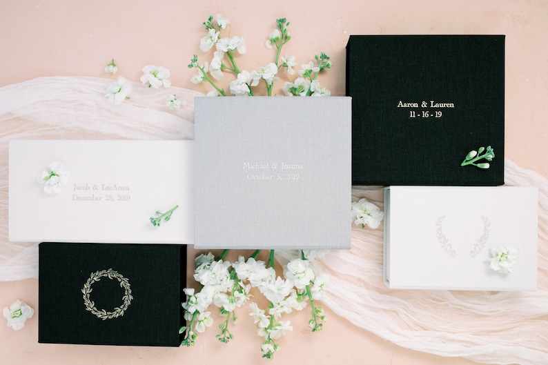 May include: A collection of five square and rectangular boxes in black, white, and light gray with fabric-like textures. Some boxes feature gold or silver foil text including names like Aaron & Lauren, Jacob & LeeAnna, and Michael & Joanna with dates.