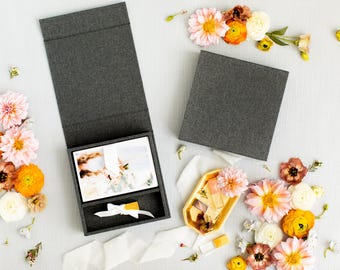 Puede incluir: Dos cajas cubiertas de tela gris oscuro, una abierta y otra cerrada, que contienen una impresión fotográfica rectangular y un pequeño objeto amarillo atado con una cinta blanca. Una bandeja hexagonal dorada contiene objetos pequeños, rodeada de flores rosas, naranjas y blancas.