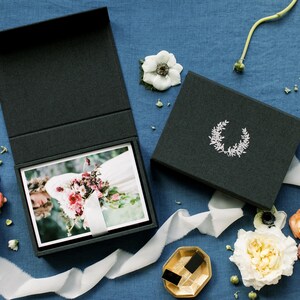 May include: A black rectangular keepsake box with a hinged lid, featuring a white laurel wreath emblem on the cover. Inside, a photograph of a floral bouquet is displayed. The box is surrounded by white silk ribbon and various fresh flowers.