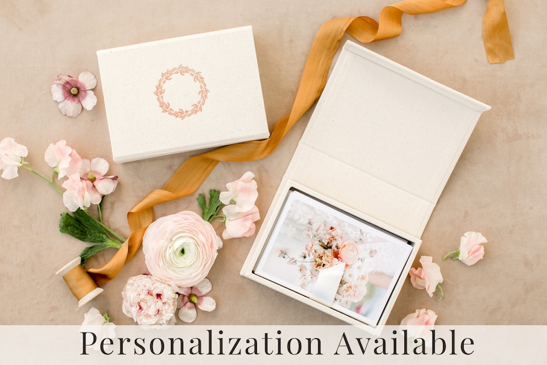May include: A cream-colored rectangular keepsake box with a copper-colored wreath emblem on the lid. The open box contains a stack of photographs. A mustard-yellow ribbon, a wooden spool, and several light pink ranunculus flowers surround the box.