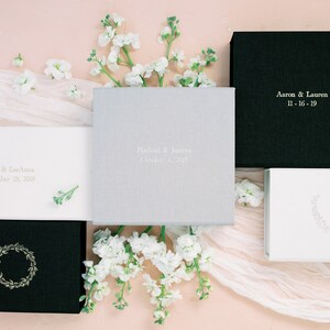 May include: A collection of five square and rectangular photo albums with fabric covers in black, white, and grey. The covers feature personalised text including names and dates, such as Aaron & Lauren 11-16-19, or decorative wreath designs.