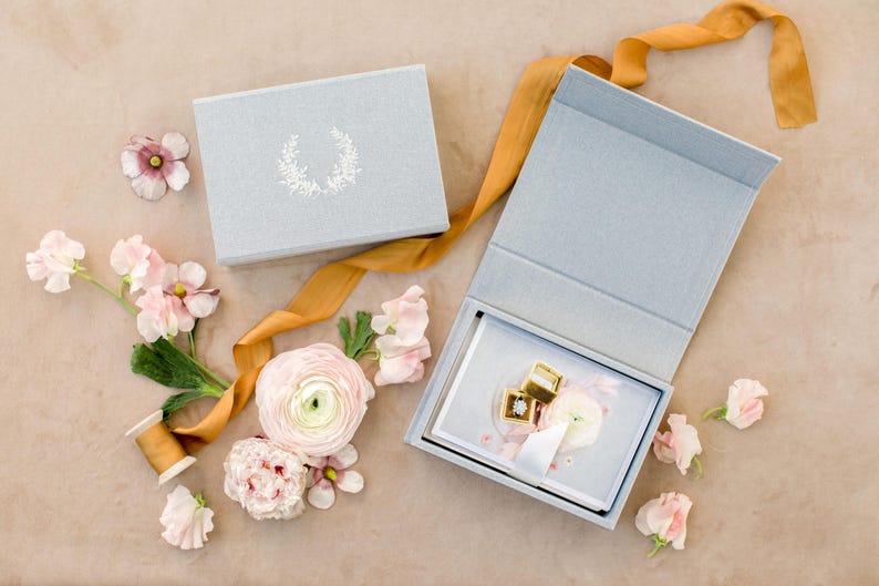 May include: A light blue rectangular box with a white floral wreath design on the lid, accompanied by an open matching box containing two gold rings and a white flower, all surrounded by loose pink ranunculus flowers and a long mustard ribbon.