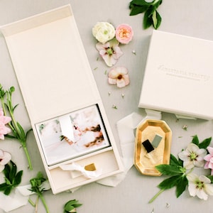 Puede incluir: Una caja de presentación crema abierta con una impresión fotográfica en el interior, junto a una caja cerrada con texto. Las cajas están rodeadas de flores, vegetación y una bandeja dorada con objetos pequeños. Una cinta está atada alrededor de la caja abierta.